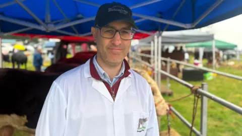 A man wearing a cap, glasses and white coat, red quarter zip and blue shirt