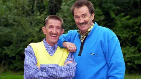 BBC Paul and Barry Chuckle smiling and looking straight on. Paul is wearing a blue and white checked long-sleeved shirt with a yellow jumper. Barry is resting his arm on his brother's shoulder and is wearing a long-sleeved blue jumper with a yellow and black shirt underneath.