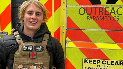 Finlay Maguire A young man with blonde hair is wearing a bullet proof vest over a black jacket. He is standing in front of one of the ambulances he has just helped to take to Ukraine. 