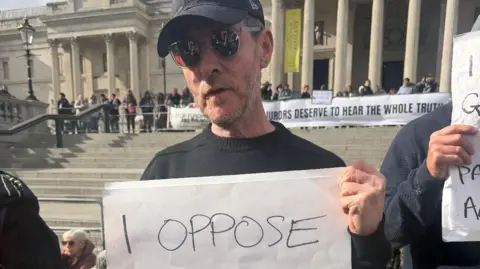 PA Robert Del Naja stands in Tralfalgar Square with his sign that reads "I oppose Genocide". He is wearing a black baseball cap, dark sunglasses and a black long-sleeved jumper.
