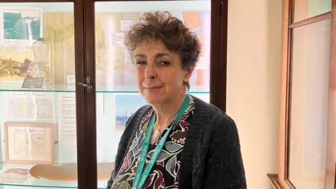 Steve Jones/BBC Elaine Hoyle stood in front of a museum cabinet of sailing maps. Elaine has short, dark curly hair and wears a grey cardigan.