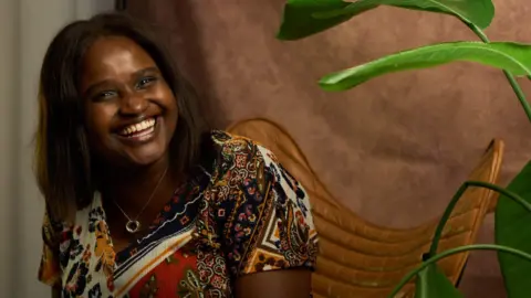 Jonny Osayamwen Louise Ndibwirende is smiling, she has shoulder length dark brown hair and wears a patterned top. There is a plant in the foreground, to one side, and she is sat on a brown leather chair with a brown fabric sheet behind her. 
