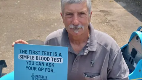 Dave Gibbons from Shottenden, Canterbury, pictured with a sign encouraging men to undertake a blood test to detect prostate cancer. Dave is wearing a grey shirt and the Prostate Cancer UK badge.