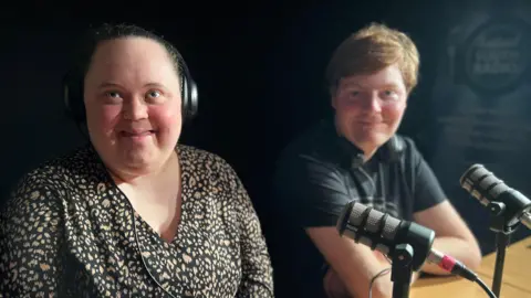 Emma Baugh/BBC Two podcasters, a young woman and a young man, sit at a desk facing the camera with a microphone in front of each of them.