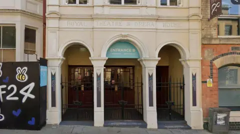 Google Neo-classical entrance to Royal Theatre, showing three cream-coloured arched doorways.The words ROYAL THEATRE AND OPERA HOUSE are engraved above the doors and one of the archways says it is ENTRANCE B. There is a hoarding to the left with F24 visible in white writing.
