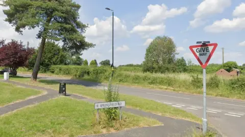 A road lined with trees and bushes. Another road, also lined with greenery, joins it at a junction.
