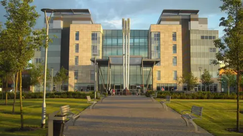 NWAFT The computer-generated image of the exterior of Peterborough City Hospital. A large glass-panelled building with a central walkway with green space on either side. The walkway has benches and is lined with trees.
