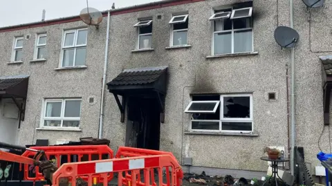 The front of a house with black smoke marks on the doorway and the windows - all windows are open, with one smashed window. There is debris on the floor outside the front of the house and an orange bollard with a forklift.