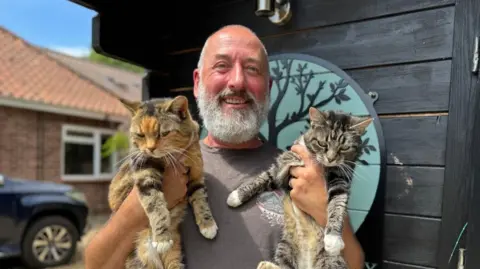 A bald men with a grey beard wearing a grey T-shirt stands holding a cat in each arm in front of a cattery