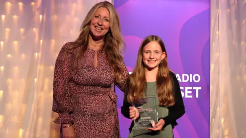 Mel Everett wearing a pink leopard patterned dress, standing beside Mia with her arm around her. Mia is wearing a green dress and a black cardigan, holding her award and smiling. In the background there is a pink BBC Radio Somerset banner and a white sheet with fairy lights underneath it.