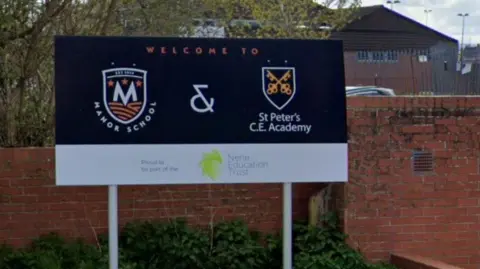 School sign at entrance for Manor School and St Peter's Academy. Both have logos on the sighn, which has a blue background and a white band at the bottom. A school building is visible behind.