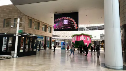 Alex Pope/BBC An indoor shopping centre walkway with several people walking. A large digital screen hangs overhead displaying colourful graphics, and a bright pink-and-green small house-like structure stands to the right.