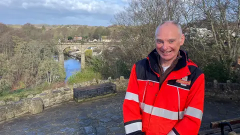BBC/Katie Radley A man in a red Royal Mail jacket is smiling at the camera, with a view of a viaduct over a river in the background with trees on either side.
