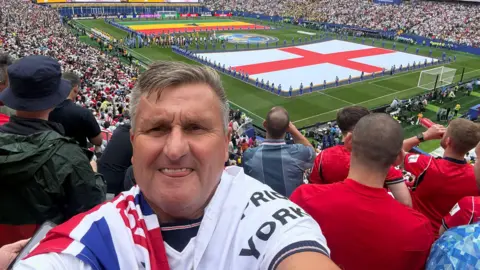 Ed Faulkner Ed Faulkner pictured ahead of kick-off at England's game against Ghana in the group stages. He is wearing a white England shirt with the flag draped over his shoulder.