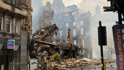The Union Corner building is reduced to rubble after a fire in Glasgow. Victorian buildings and street furniture surround the iron and sandstone remains. 