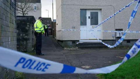 PA Media Police cordon off the scene in Harborne Drive, West Calder. An officer wearing a high-vis jacket can be seen in the garden. The house with the white door is brown. In the foreground, there is white and blue police tape.