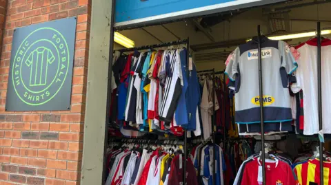 An open shop entrance displays racks of football shirts inside, with a sign reading “Historic Football Shirts” on the brick wall outside.
