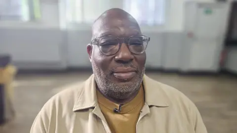 The photo shows a black man with a bald head and grey stubble. He is wearing grey rimmed glasses and looking directly at the camera. He is wearing a mustard t-shirt under a cream overshirt.