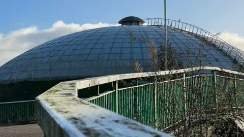 A view of the large built dome from a railing - both look like they need cleaning, with foliage growing. Under a blue sky.