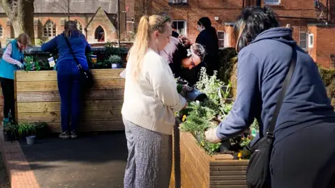 Northampton BID A group of people, placing plants into two large wooden planters. They are mostly leaning over. A church and a number of buildings are behind them.