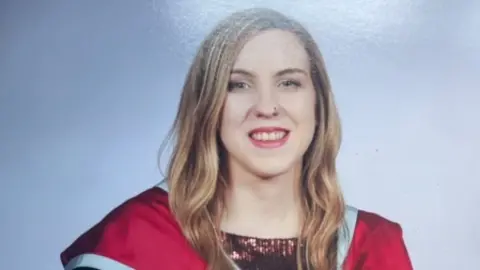 Family handout A woman with long fair hair looks into the camera. She is smiling, She has nose ring. She's wearing a red graduation gown with a light blue trim and a wine coloured sparkly top is visible underneath