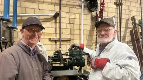Two men in caps and glasses, one has a grey beard. They are both looking directly at the camera and smiling. They are stood either side of a piece of machinery