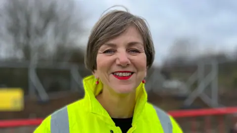 Lillian Greenwood is smiling at the camera. She's wearing a hi-vis jacket and red earrings. Behind her we can see construction work is taking place.