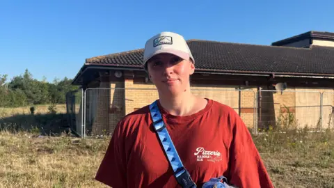 Holly Nichols/BBC Laura Watson-Smith standing in a field with a boarded up single-storey yellow brick building behind her. She is wearing a white baseball cap and a red T-shirt.