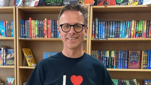 A man with short brown hair, wearing black rimmed glasses and a blue sweater with "I heart" written on it, standing in front of bookshelves filled with colourful books.