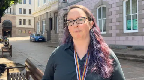 Louise Doublet is standing outdoors on a sunny day, in front of a building with light pink and grey stonework. She has long, wavy, purple-tinted hair and is wearing glasses and a dark-coloured shirt. Around her neck is a rainbow-striped lanyard.