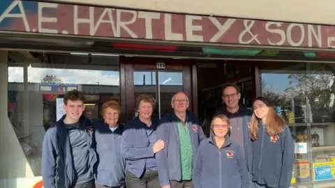 Simon Thake/BBC A group shot of people of all ages in matching navy fleeces outside a shop.