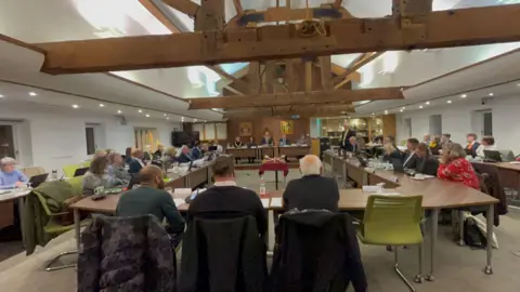 The photo shows the council chamber which has a high ceiling featuring exposed wooden beams. The councillors are seated around a U-shaped arrangement of tables, in a council meeting. Each is equipped with papers, laptops, and drinks. 