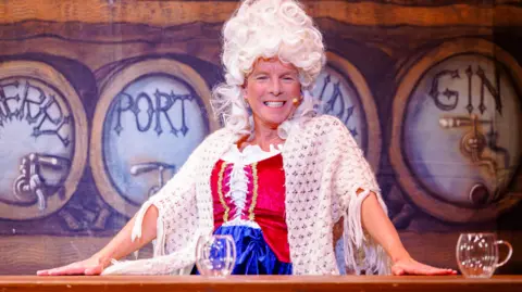 Fourth Wall Photography Terry, dressed in a white wig, with a microphone on his face, wearing a white top, bodice, blue skirt and shawl. He is at a pretend bar, with two glasses in front of him. 