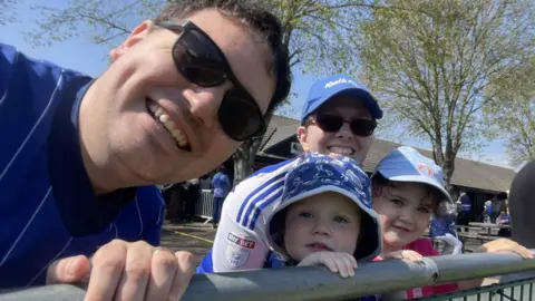 Contributed Sam Hall takes a selfie with his two young daughters and his partner at the Martello Ground during a Town match. He has short dark hair and wears sunglasses as well as a Town shirt.