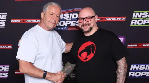 Supplied American wrestler Bret Hart wearing a white top and shaking hands with Dante Richardson who is wearing a black top with a red and black logo. 