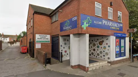 Google A red brick building with two blue banners reading Allcare Pharmacy and Travel Clinic on either side of the building in view in the picture