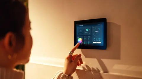A female is adjusting lighting and room somesthesia pinch a smart location system. She's rubbing a mini surface mounted to a wall. She's wearing a beige turtle cervix jumper. 
