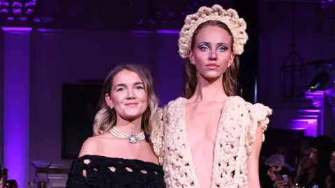 Getty Images Shonagh Murray (left) with a model wearing one of her designs. Shonagh has long blonde hair which has been curled and wears a black knitted off-the-shoulder dress and a pearl Vivienne Westwood necklace. The model on the right is considerably taller than Shonagh and wears a low-cut cream knitted dress with a matching crown-style headband. The stage behind them is lit purple. 