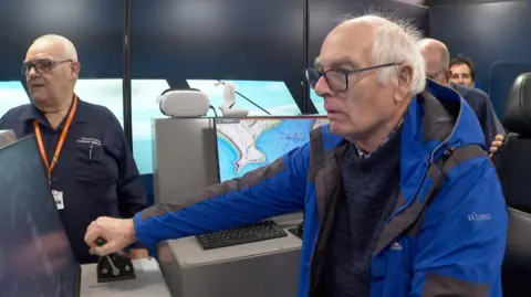 Retired fishermen Colin Warwick trying to steer a boat in the navigational simulator at the Marine Academy in Kingswear. He is wearing a blue and black coat and has white hair and black glasses. Fishing maps are behind him.