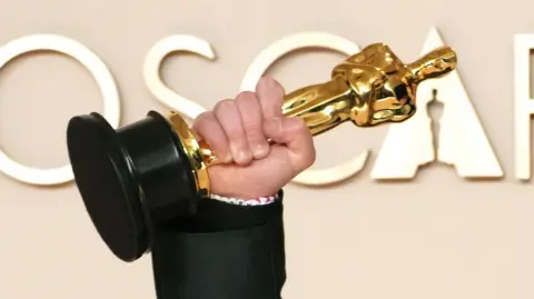 A close-up of Kieran Culkin's hand holding his Oscar statuette aloft in front of the word "Oscars" in large lettering behind him at the 2025 ceremony