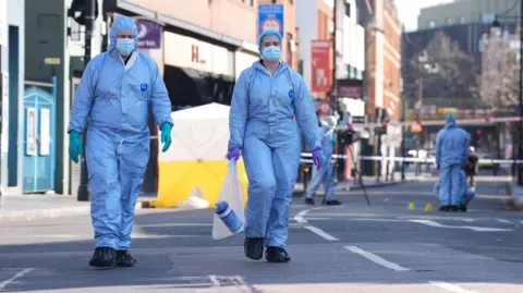 PA Media Two forensic officers dressed in blue hazmat suits walk towards the camera in front of a white and yellow police tent and cordon