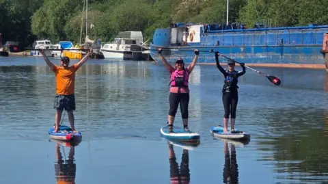 Natasha Sones Three people on stand up paddle boards in a dock. One is a man in an orange top, then a woman in pink and another woman in black. They are all holding paddles above their head and standing on stand up paddle boards 
