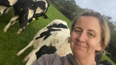 Marie Stephenson, woman with blonde hair smiling. She is in a field with several cows in the background with her.