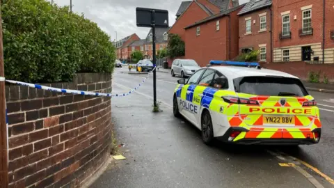 A section of blue and white police tape cordons off a length of pavement, flanked by a police car
