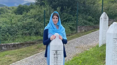 Una Srabovic-Ryan Una is standing next to her father's grave stone. She is wearing a blue dress and a blue hijab.