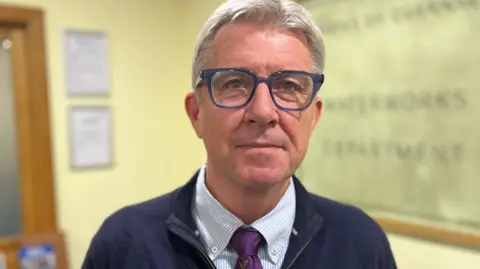 Deputy Mark Helyar - A man with grey hair in a small quiff, wearing blue rimmed glasses. He is wearing a blue jacket, with a blue and white checked shirt, with a purple tie. 