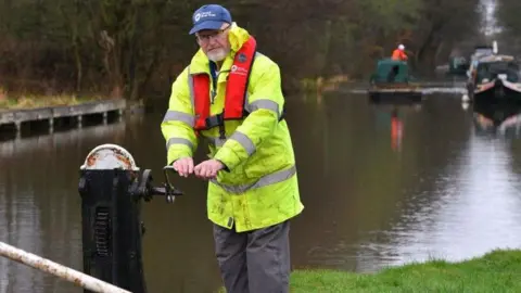 Canal & River Trust A man in a yellow hi vis jacket, red life jacket and blue cap, operating a lock by a canal 