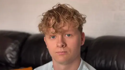 A head and shoulders of Harry Pyett, who has curly light brown hair and blue eyes, and is looking at the camera. He is sitting on a dark-coloured leather sofa indoors.