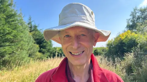 Tom Mackenzie is wearing a red collared top, and a cream bucket hat, smiling at the camera with trees, hedges and a blue sky behind him.