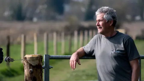 Herefordshire Wildlife Trust Hutton, wearing a grey t-shirt, leans on a metal gate and looks off into the distance.